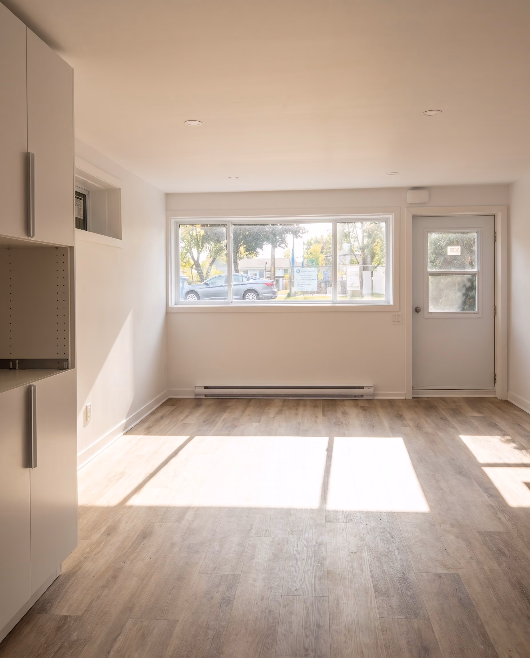 Pièce vide fraîchement rénovée avec une grande fenêtre et une porte vitrée, plancher de bois clair.